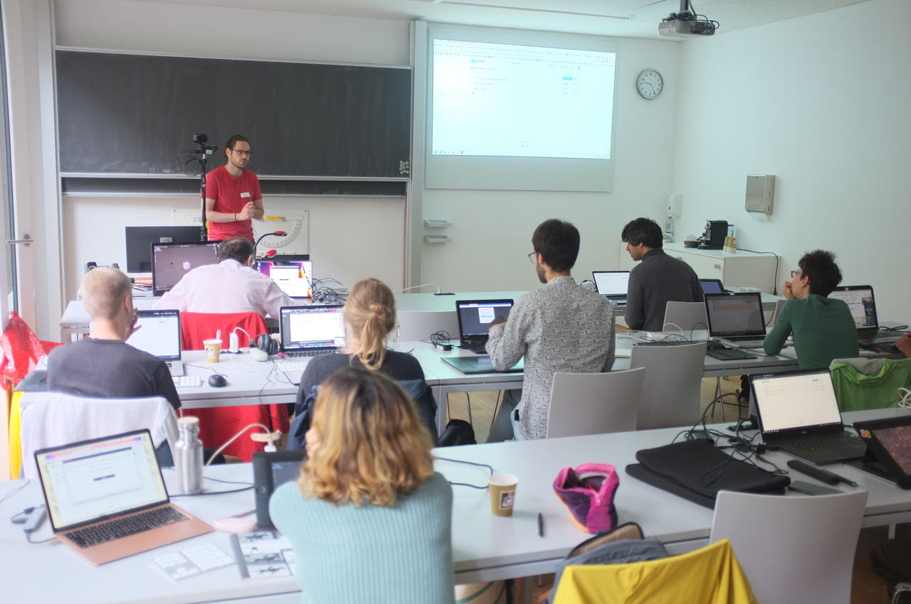 Professional trainer conducting a Blender 3D software workshop for a group of attentive students in a modern classroom setting. The instructor is demonstrating advanced 3D modeling techniques on a large screen, while participants follow along on their laptops. The room is well-lit with natural light, and the atmosphere is focused and collaborative, highlighting the hands-on learning experience.
