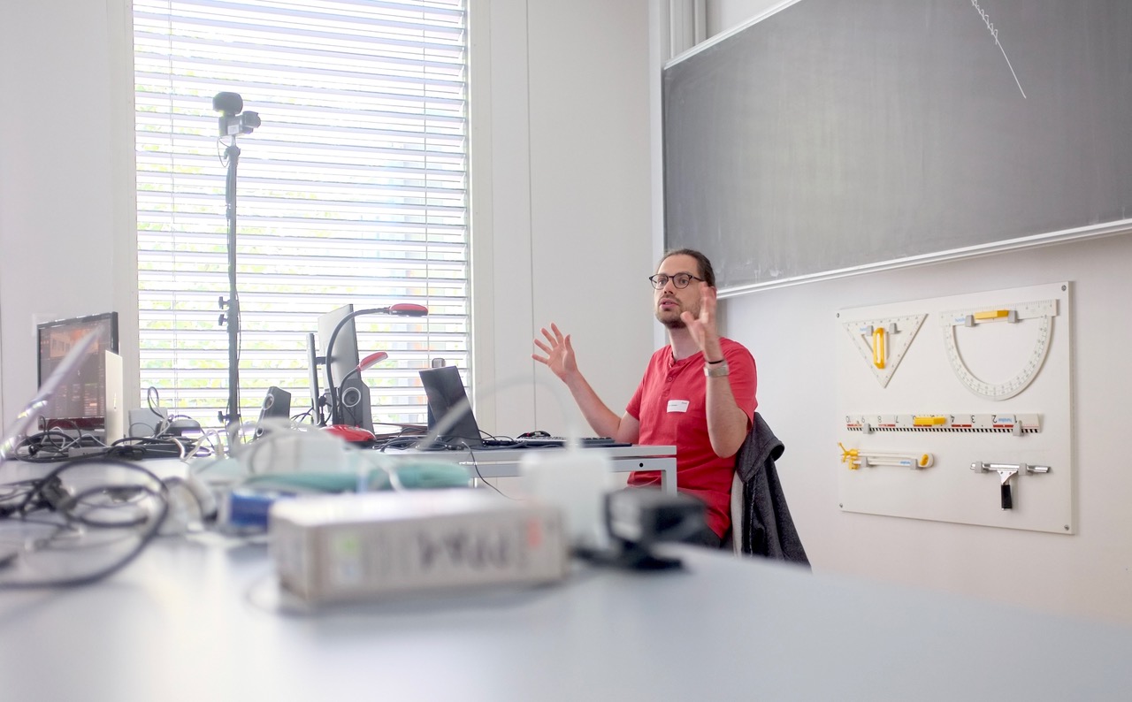 Professional trainer working with students at a computer workstation. Both are engaged in learning Blender 3D software, with the trainer pointing at specific elements on the screen while providing individualized instruction. The setting appears to be a modern office space with natural lighting, creating a focused and collaborative learning environment.